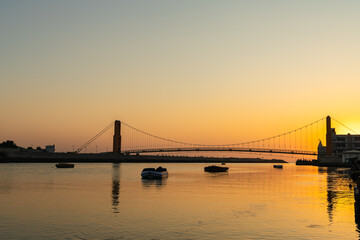 Obraz premium silhouette of suspension bridge and boats during golden sunset over river
