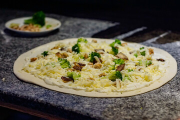 Raw white-sauce pizza topped with broccoli, mushrooms, and cheese on a stone counter, showing the artisan cooking process, fresh ingredients, and homemade preparation in a kitchen environment