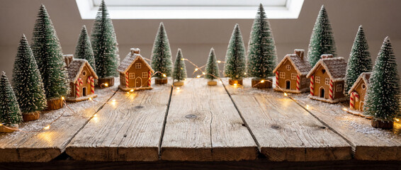 Snowy gingerbread village with christmas trees and fairy lights arranged on rustic wooden table beneath bright window in cozy winter setting