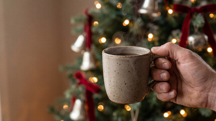 Hand holding a ceramic mug of hot beverage in front of a decorated christmas tree with festive ornaments and glowing string lights
