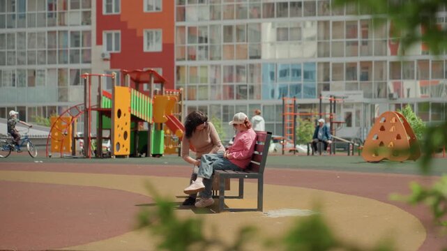 Caucasian Mentor Approaches Teen On Bench At Playground, Colorful Slide And Highrise Apartments In Background, Encouraging Smile And Guided Conversation, Supportive Urban Courtyard Scene