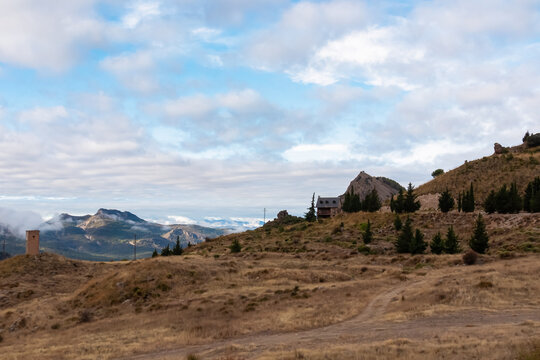 A solitary stone refuge stands on a grassy hill beside a winding dirt path, overlooking the misty peaks of Sierra Nevada, Andalusia, offering a scenic hiking route in the Spanish mountains.