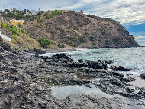 Jagged volcanic rocks and tide pools line the foreground, leading toward the turquoise sea and the historic Torre del Diablo on a rugged hill in Almunecar, capturing the wild beauty of coastal Spain. - Powered by Adobe