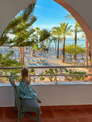 Obraz premium A little girl sits on a balcony chair gazing at the sandy beach and palm trees of a luxury resort in Almunecar. The child enjoys the ocean view through the terrace railing during a sunny holiday.