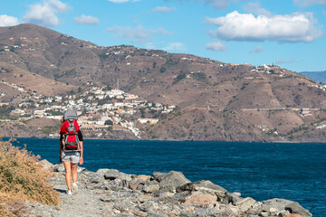 A woman carrying a baby in a red backpack hikes along a rocky coastal path near the blue sea in Almuñécar, enjoying the scenic views of the white hillside town on the Costa Granadina in Spain.