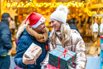 Smiling couple in a Bavarian Christmas market holding wrapped gifts surrounded by winter magic and lights