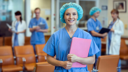 Confident smiling nurse in blue scrubs holds pink folder surrounded by hospital team members