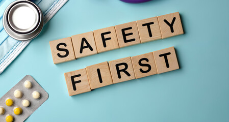 Wooden blocks spell SAFETY FIRST near stethoscope pills and face mask. Medical instruments are lying on a pastel blue surface. Healthcare concept for insurance and emergency.