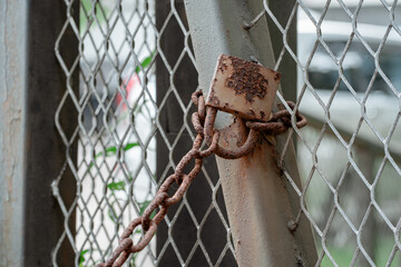 An old square rusty padlock and corroded chain locking a galvanized chain-link fence gate.