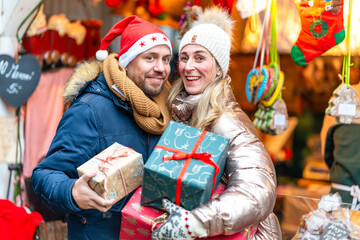 Smiling couple holding gifts at a Bavarian Christmas market filled with lights festive colors and winter joy