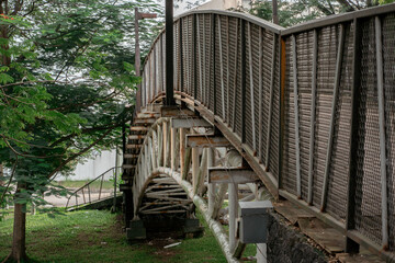 Entrance ramp of a metal pedestrian bridge with safety railings surrounded by green tropical trees.
