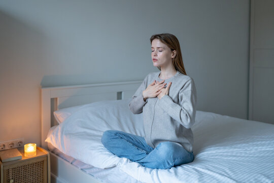 Calm woman practicing mindful breathing sitting on bed at home, hands on chest, eyes closed. Emotional self-regulation, stress relief, anxiety management, inner balance, body awareness, meditating. 