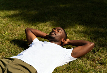 A young Black man relaxing on the grass with his eyes closed. Resting outdoors in a park on a sunny summer day. Wellness and mental health concept