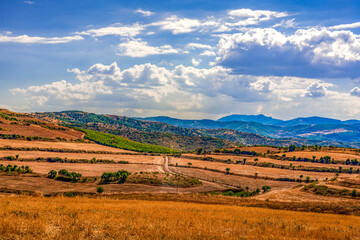 Naklejka premium Bright Summer Landscape with Golden Fields, Rolling Terraced Hills, and Distant Blue Mountains Under a Sky of Sunlit Clouds