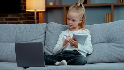 Caucasian clever pupil kid child girl daughter concentrated learn studying with e-learning web course on couch writing notes raising hand answer to teacher study learning with laptop at evening home