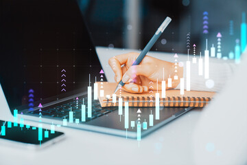 Person writing in notebook over laptop with financial forex chart graph overlay on digital screen background, symbolizing market analysis and growth.