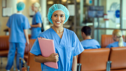 Confident smiling nurse in blue scrubs holding patient charts in busy hospital corridor setting