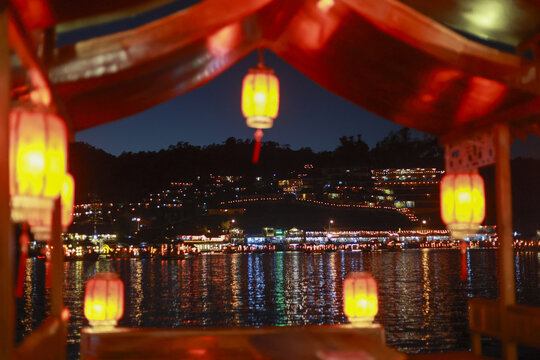 Serene romantic night view from inside traditional Chinese boat with glowing red lantern. city light and scenic landscape create beautiful reflection on water - Powered by Adobe