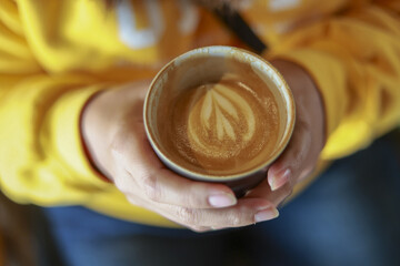 Close up of hand holding warm cup of coffee with beautiful latte art. cozy person wearing yellow sweater enjoying fresh and delicious morning drink with caffeine