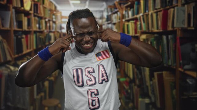 Man with fingers in ears grimacing in library wearing usa jersey and wristbands; study distraction frustration.