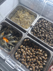 Fresh seafood display at food market featuring various raw ingredients. An appetizing assortment of clam, shrimp, and sea snail in steel containers, ready for cooking