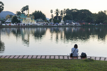 Solitary person, woman alone, sitting with her back to camera, looking at view. calm lake water reflects beautiful temple during peaceful and serene sunset