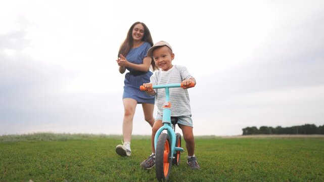 A boy rides a blue bike on the grass while a woman follows closely. The boy maneuvers the blue bike forward as the woman runs on the open grass field.
