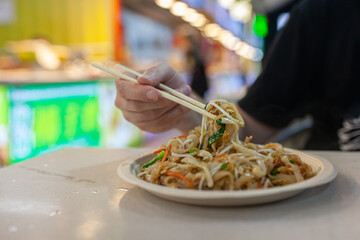 Close-up of paper plate of stir-fried noodles with egg and vegetables lifted by chopsticks. Casual street-food moment in night market in Sanya, Hainan. Local cuisine and travel culture concept.