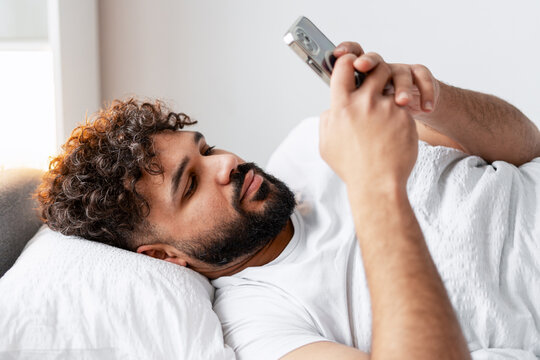 Young man relaxing in bed, checking smartphone for news and social media content
