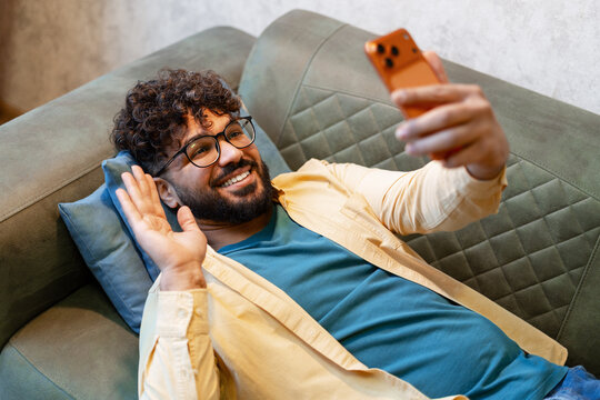 Young man relaxing on couch, connecting with a video call, smiling and waving - Powered by Adobe