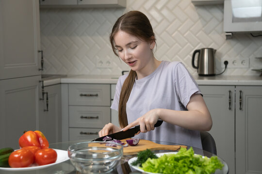 Young girl is carefully cutting a red onion on a wooden cutting board in a modern kitchen, surrounded by fresh vegetables like tomatoes, cucumbers, lettuce, and dill, preparing a healthy meal - Powered by Adobe