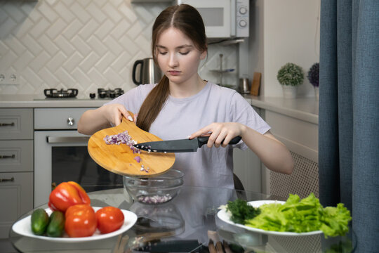 Young girl pouring sliced red onion into bowl in modern kitchen surrounded by fresh vegetables such as tomatoes, cucumbers and lettuce, highlighting healthy cooking and lifestyle - Powered by Adobe