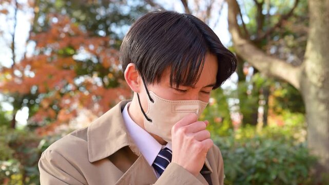 秋冬の紅葉の屋外で風邪をひいてマスクし、寒気や発熱や頭痛を感じ咳き込むコートを着たビジネスマンの動画