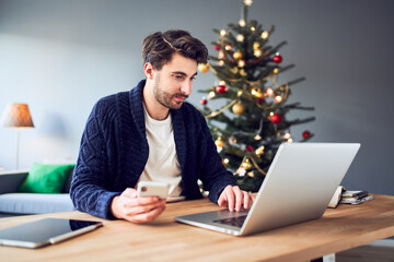 Man paying with mobile phone for Christmas shopping using laptop at home