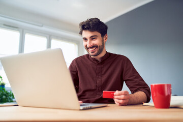 Cheerful young man shopping online using laptop and credit card