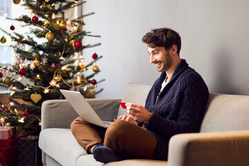 Young man sitting on sofa shopping for Christmas presents online paying with credit card