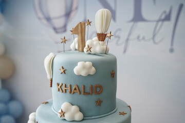 A soft blue two-tier first birthday cake decorated with clouds, hot air balloons, and gold accents. A stylish and whimsical cake design perfect for baby celebrations, party styling, and milestone even