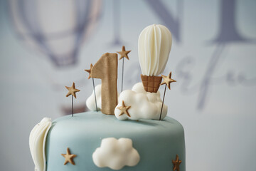 Close-up detail of a pastel blue first birthday cake featuring a gold number one, hot air balloon topper, fluffy cloud decorations, and star accents. A soft, elegant design perfect for baby celebratio