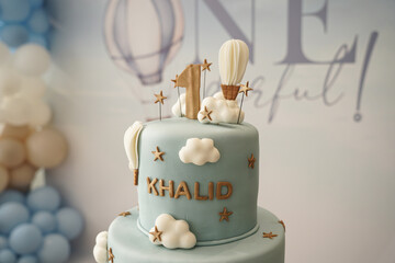 A soft blue two-tier first birthday cake decorated with clouds, hot air balloons, and gold accents. A stylish and whimsical cake design perfect for baby celebrations, party styling, and milestone even