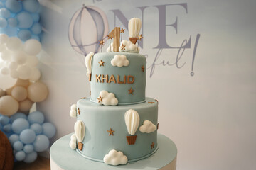 A soft blue two-tier first birthday cake decorated with clouds, hot air balloons, and gold accents. A stylish and whimsical cake design perfect for baby celebrations, party styling, and milestone even