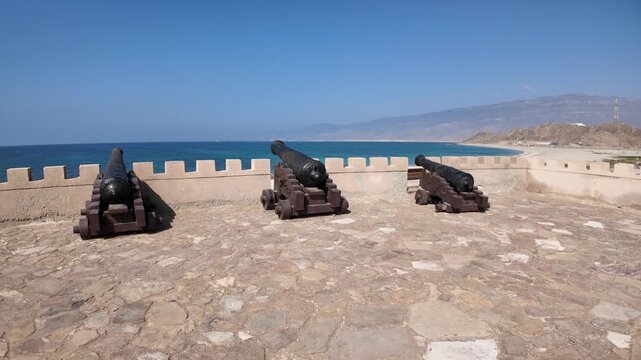 Ancient cannons positioned on the historic Mirbat fort wall, overlooking the pristine sandy beach and turquoise waters of the Arabian Sea in Dhofar, Southern Oman, showing coastal defense in Mirbat
