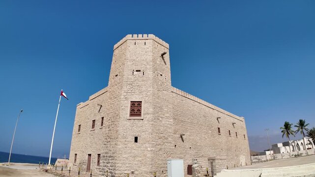 Mirbat fort. A historical stone structure with battlements and corner towers. Stands under a clear blue sky. Proudly flying the Omani flag. Representing national heritage and strength in Dhofar region