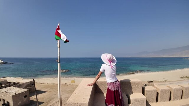 Woman on the fortified walls of Mirbat Fortress, overlooking Mirbat city, mosque minaret, Omani flag, sandy beach, and the clear blue Arabian Sea under a sunny sky in Dhofar in Oman