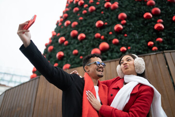 Happy diverse couple taking a selfie in madrid, celebrating christmas holidays with a festive decorated tree