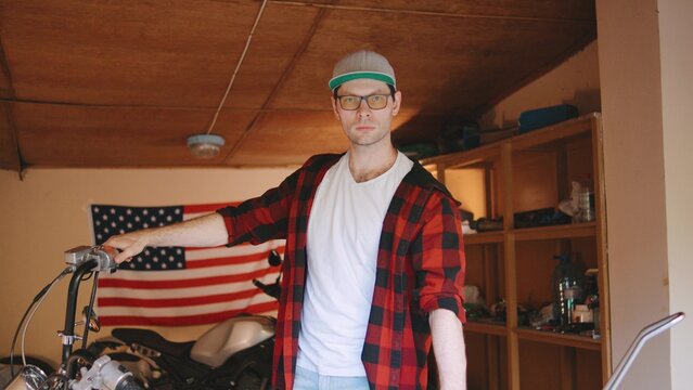 Portrait of biker man in a checkered shirt working on a motorcycle in a garage decorated with an American flag in the backdrop during daylight - Powered by Adobe