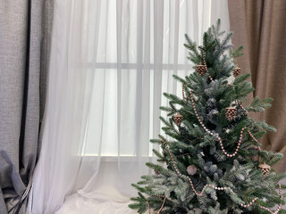 christmas tree in the window. A modern white matte Christmas tree topped with rose gold garland and glittery pine cones sits against a window with sheer white curtains and taupe drapes.