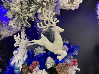 Close-up of a Christmas tree branch with a white beaded reindeer ornament, snow-covered pine needles, blue tinsel, red berries, and a large snowflake, all under blue lighting