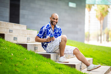 Smiling man in blue shirt and shorts sitting on outdoor steps using smartphone on a sunny day