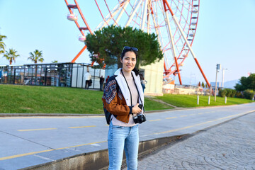 Naklejka premium Camera in hand, traveler explores outdoor amusement park by a towering ferris wheel, casual fashion, confident stance, daytime scene, photography subject capturing joyful leisure and urban scenery