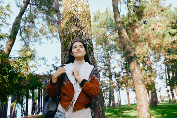 Naklejka premium Woman leaning against a tree in a sunlit park, wearing a warm jacket and scarf, enjoying nature among tall pines for a calm outdoor moment blending fashion and forest atmosphere